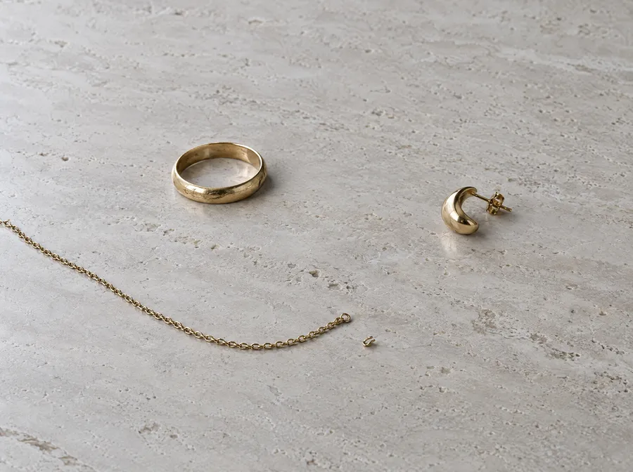 Drei Stücke Altgold mit viel Abstand auf hellem Travertin: ein getragener Ehering, eine gerissene feine Kette und ein einzelner Ohrring
