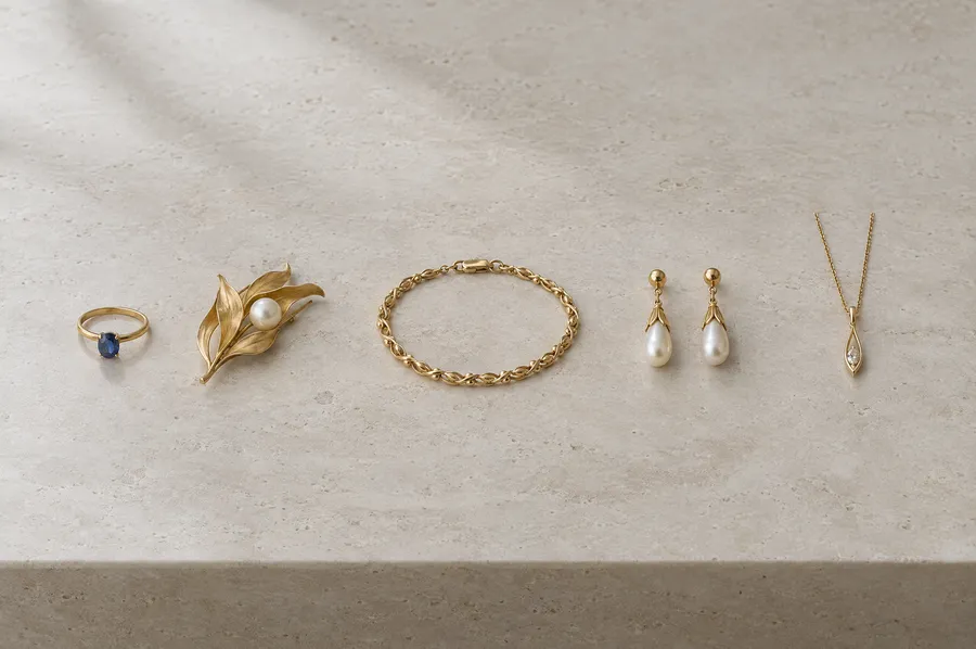 Fünf alte Goldschmuckstücke in einer Reihe auf hellem Travertin: ein Saphirring, eine Perlbrosche, ein Armband, Ohrhänger und ein Anhänger